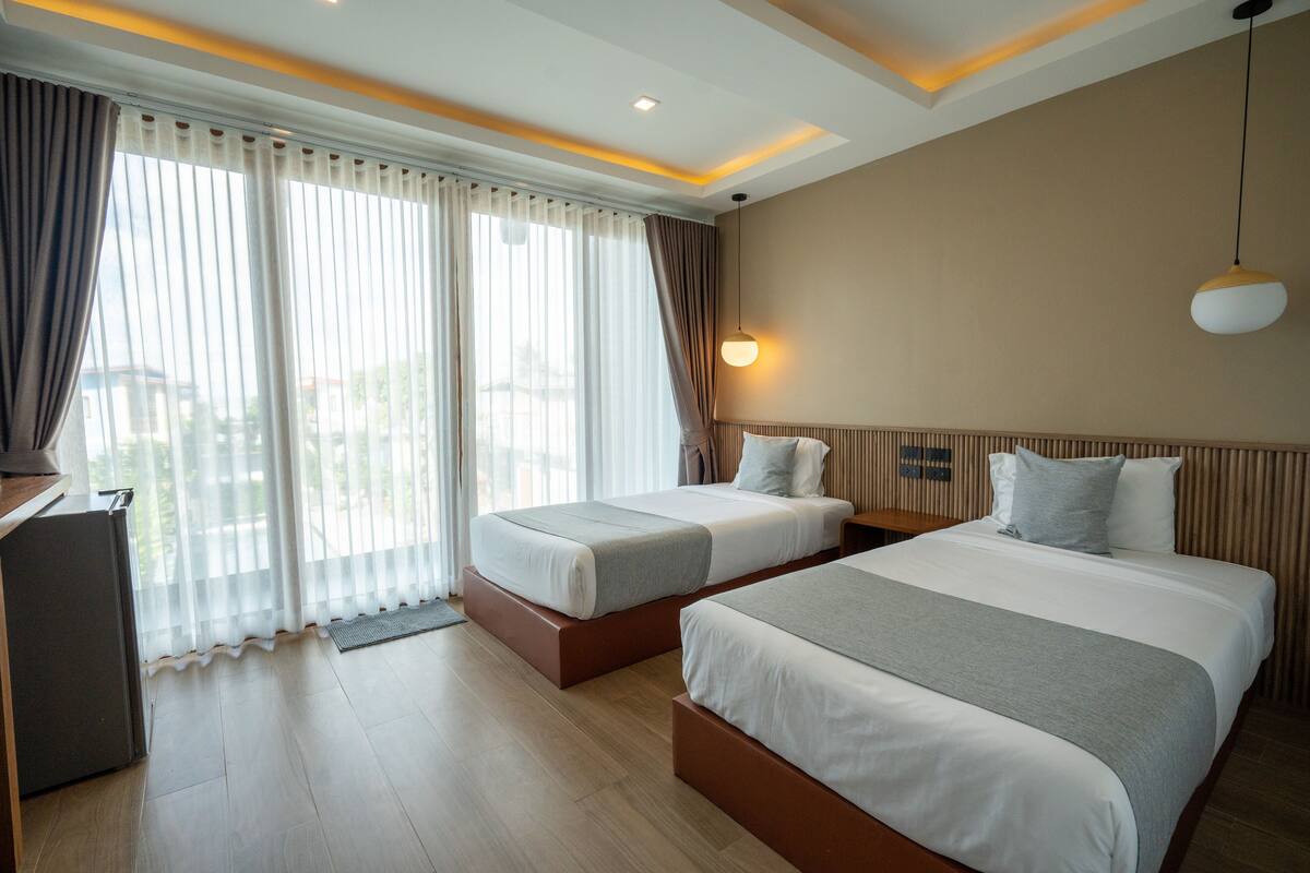A hotel room with two beds, a desk, and a view through large windows.