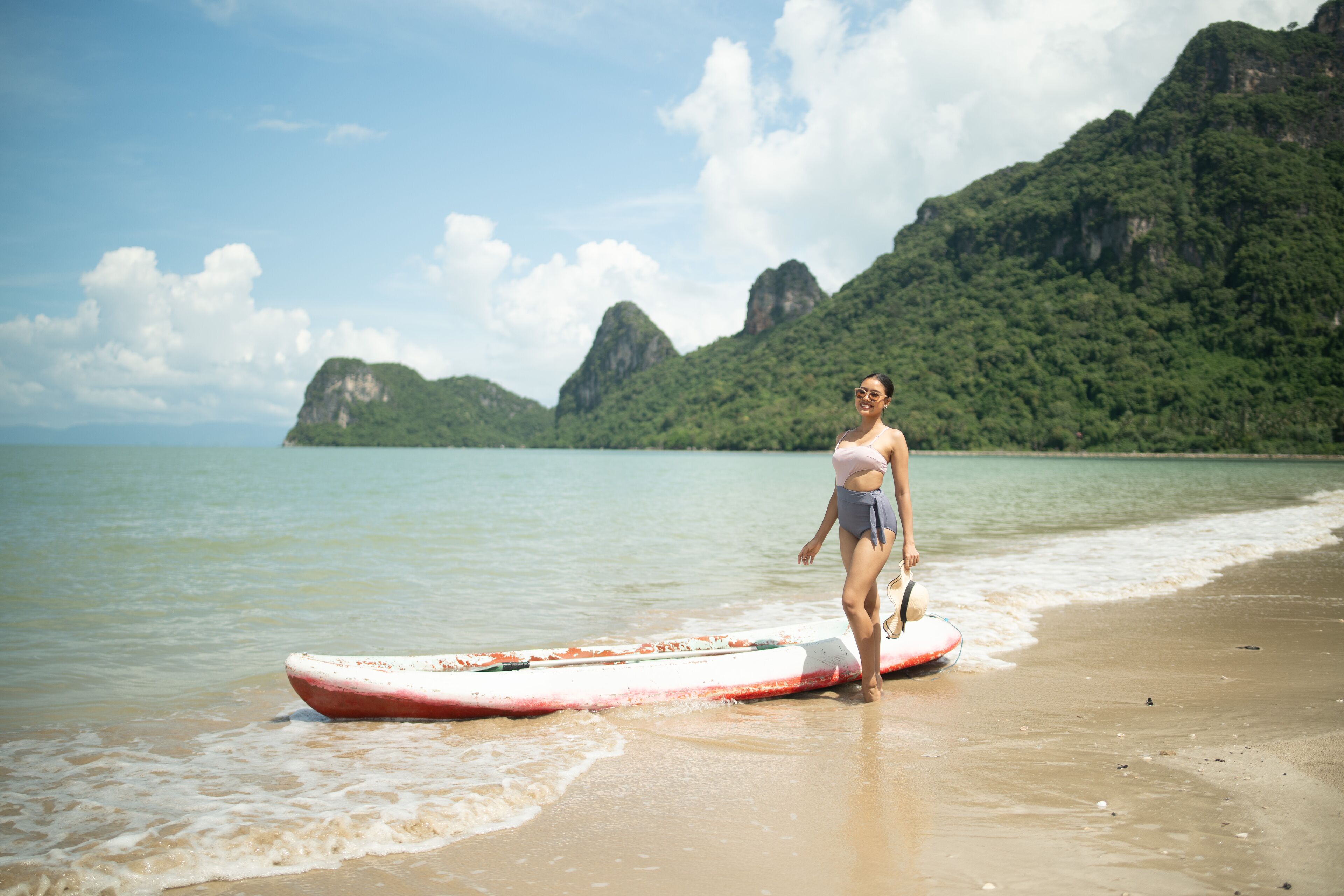 A person standing on a beach with a surfboard, surrounded by water and green hills in the background.