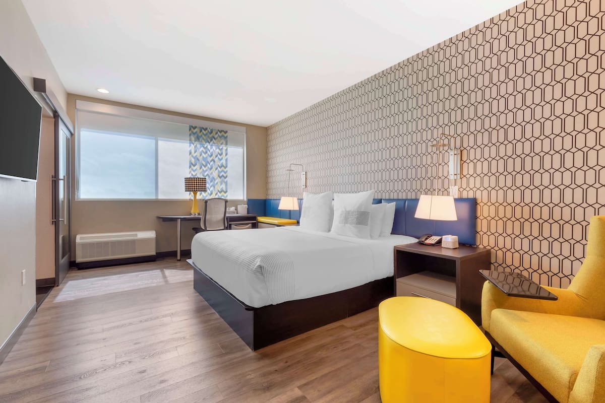 A modern hotel room with a large bed, a desk, a chair, and a window with curtains.