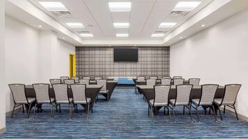 A conference room with a blue patterned carpet, rows of chairs, a large flat-screen TV, and a yellow door.