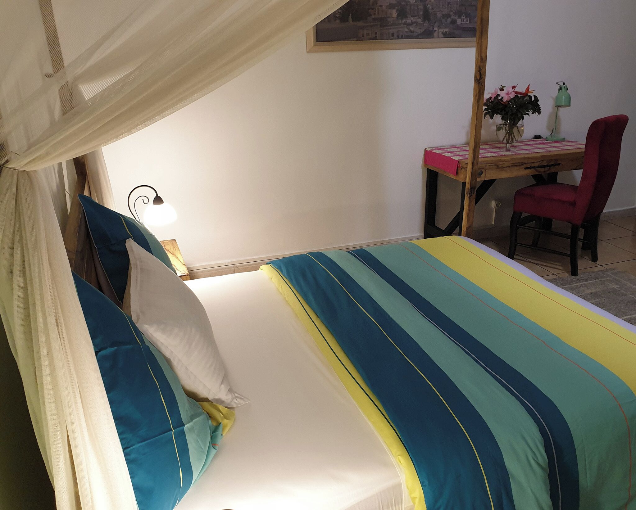 Een bed met een gestreept laken, een bedlampje, een bureau met een stoel, en een plant.
