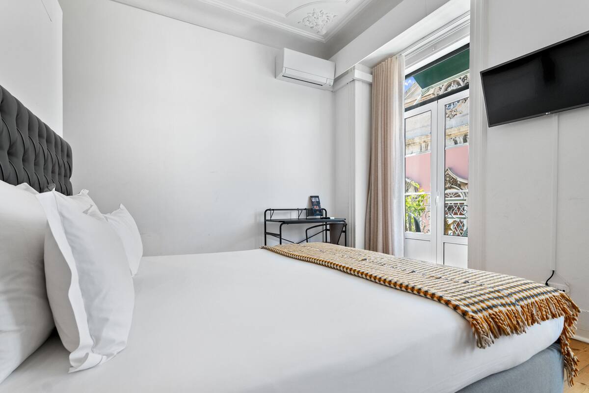 A hotel room with a bed, a desk, an air conditioner, a TV, and a window with a view of buildings.
