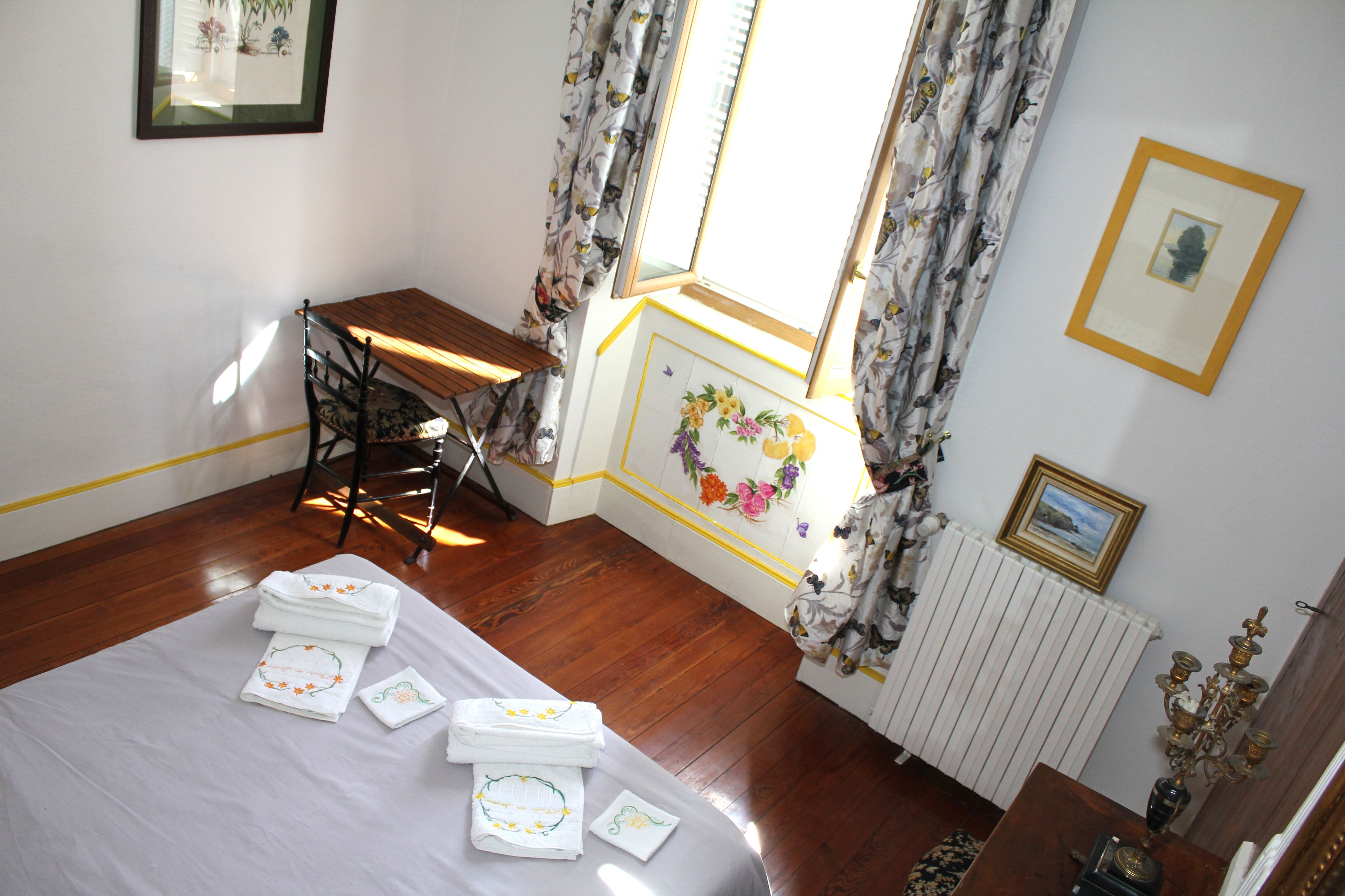 Una habitación con piso de madera, una cama con toallas ordenadas, una silla, una mesa y una ventana con cortinas.