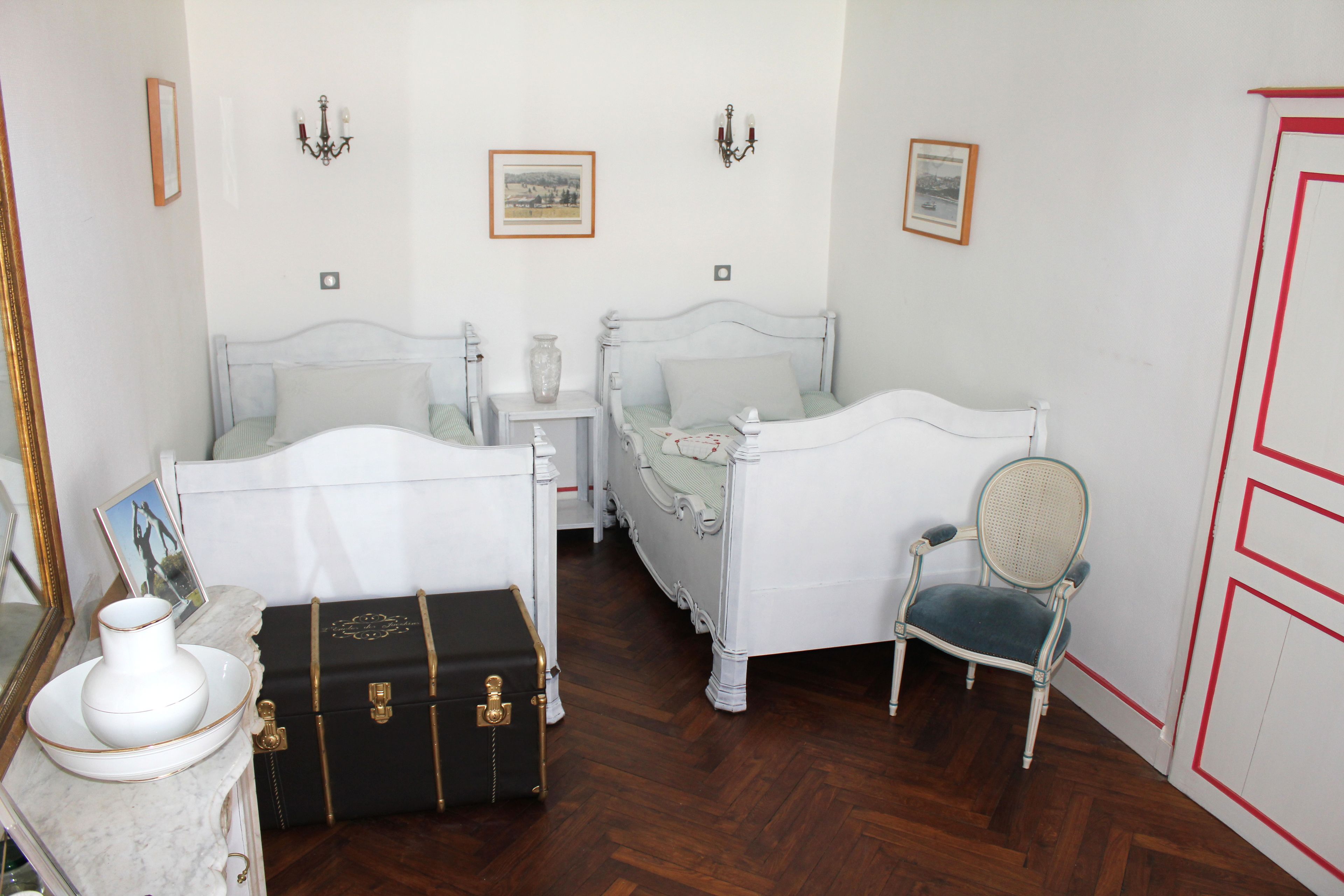Une pièce avec deux lits blancs, une valise ancienne, une chaise et une table en marbre sur laquelle est posé un vase.