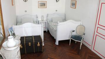 Une pièce avec deux lits blancs, une valise ancienne, une chaise et une table en marbre sur laquelle est posé un vase.