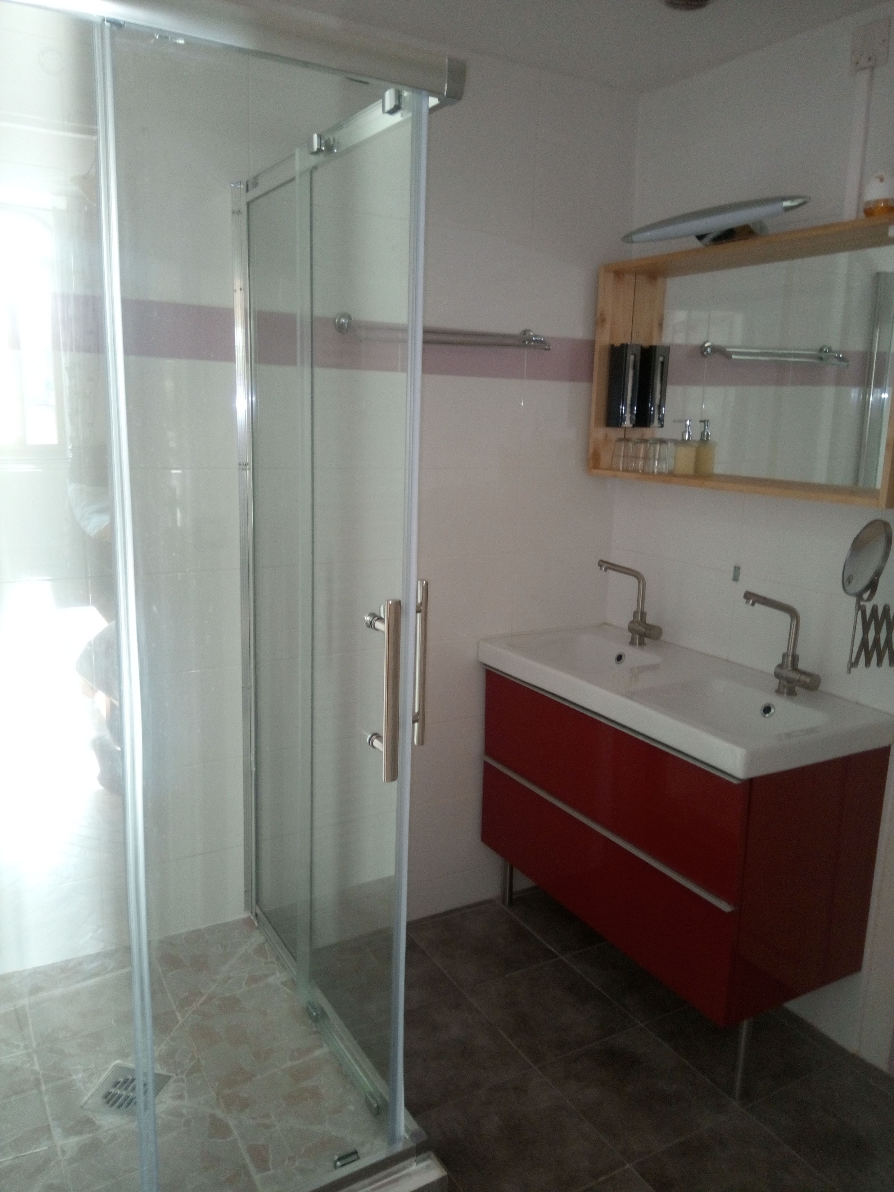 Un baño con mampara de vidrio, un lavabo blanco con tocador rojo y un espejo sobre el lavabo.
