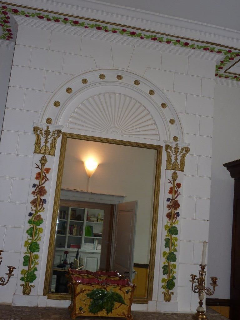 Une pièce dotée d’un grand miroir encadré de motifs dorés et de décorations florales. Une petite table surmontée d’un plateau décoratif se trouve au premier plan.