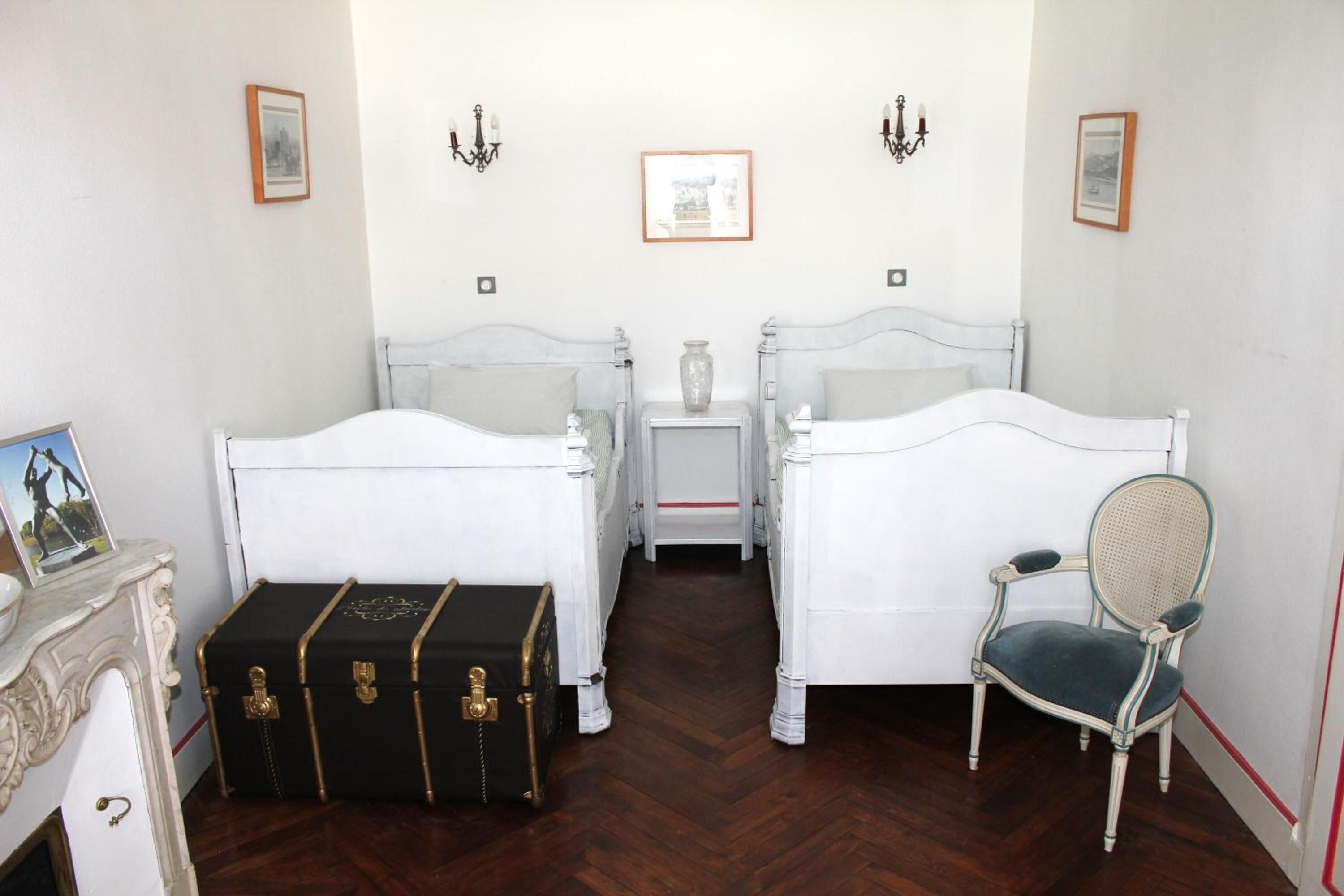 A room with two white beds, a small table with a vase, a chair, and a black trunk with gold accents.