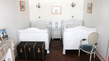 A room with two white beds, a small table with a vase, a chair, and a black trunk with gold accents.