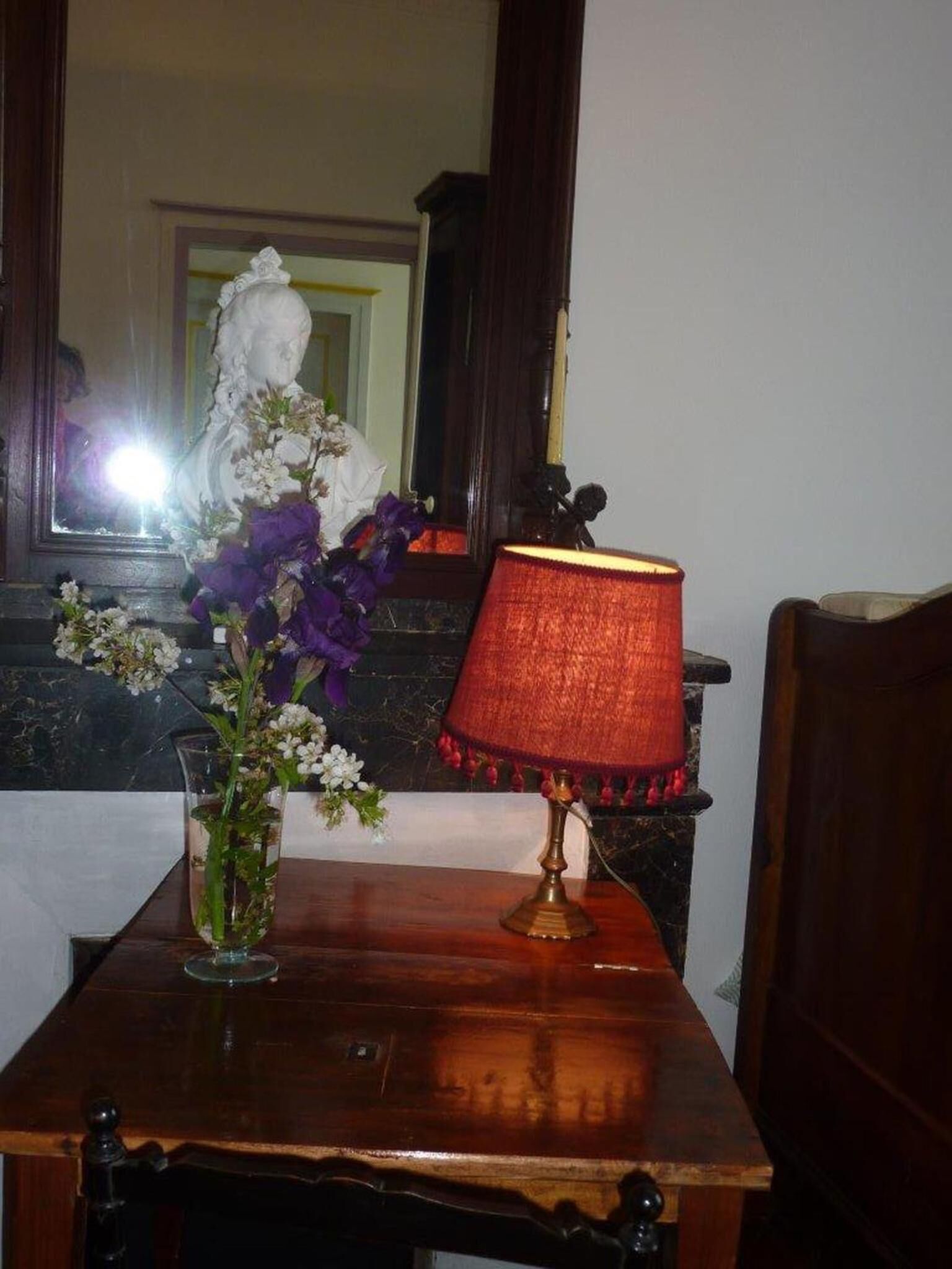 Une table en bois, une lampe rouge, un vase rempli de fleurs et un miroir qui reflète une statue.