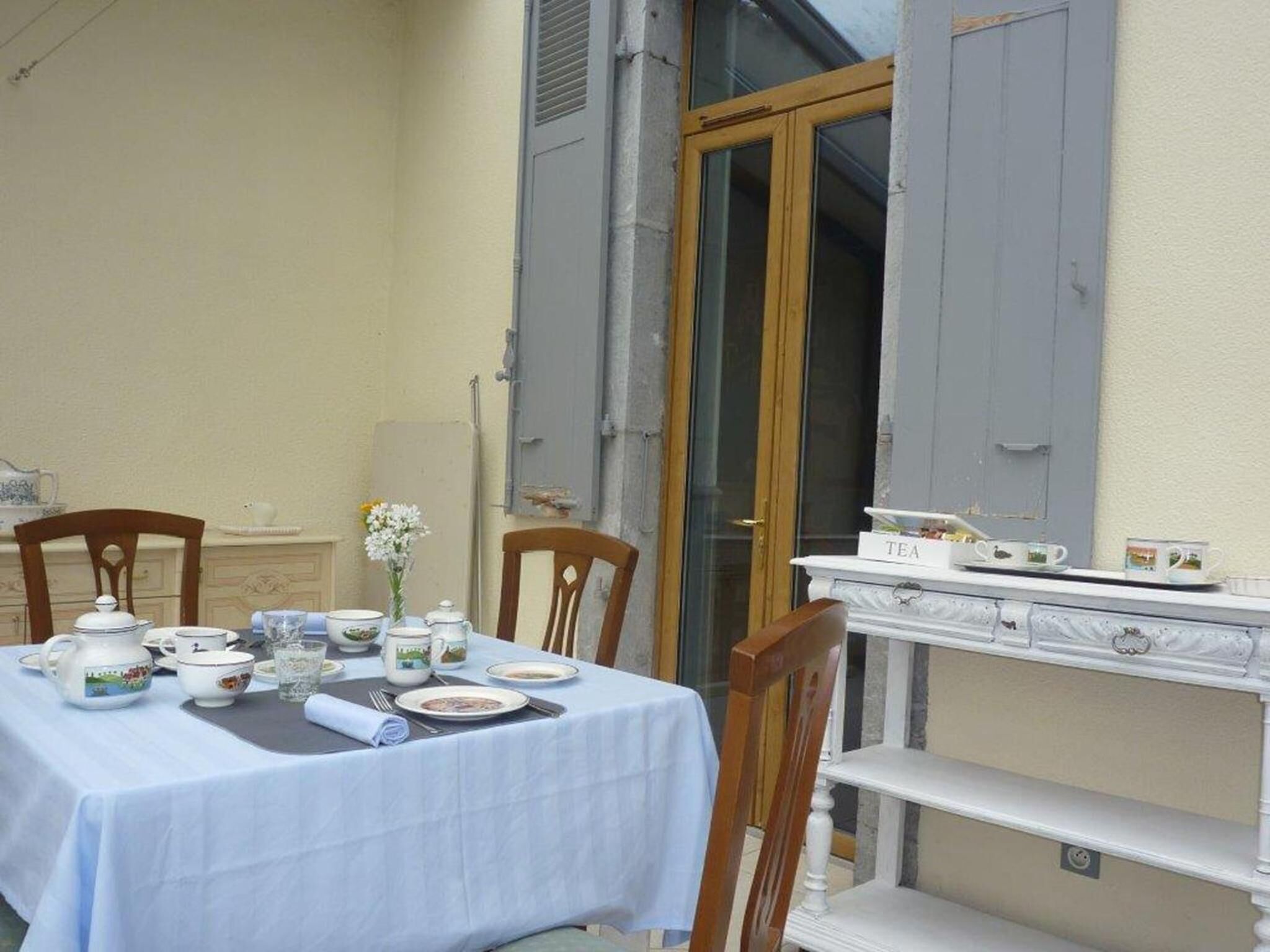 Una mesa preparada para tomar té, con mantel blanco, sillas de madera y un aparador blanco con vajillas de té.