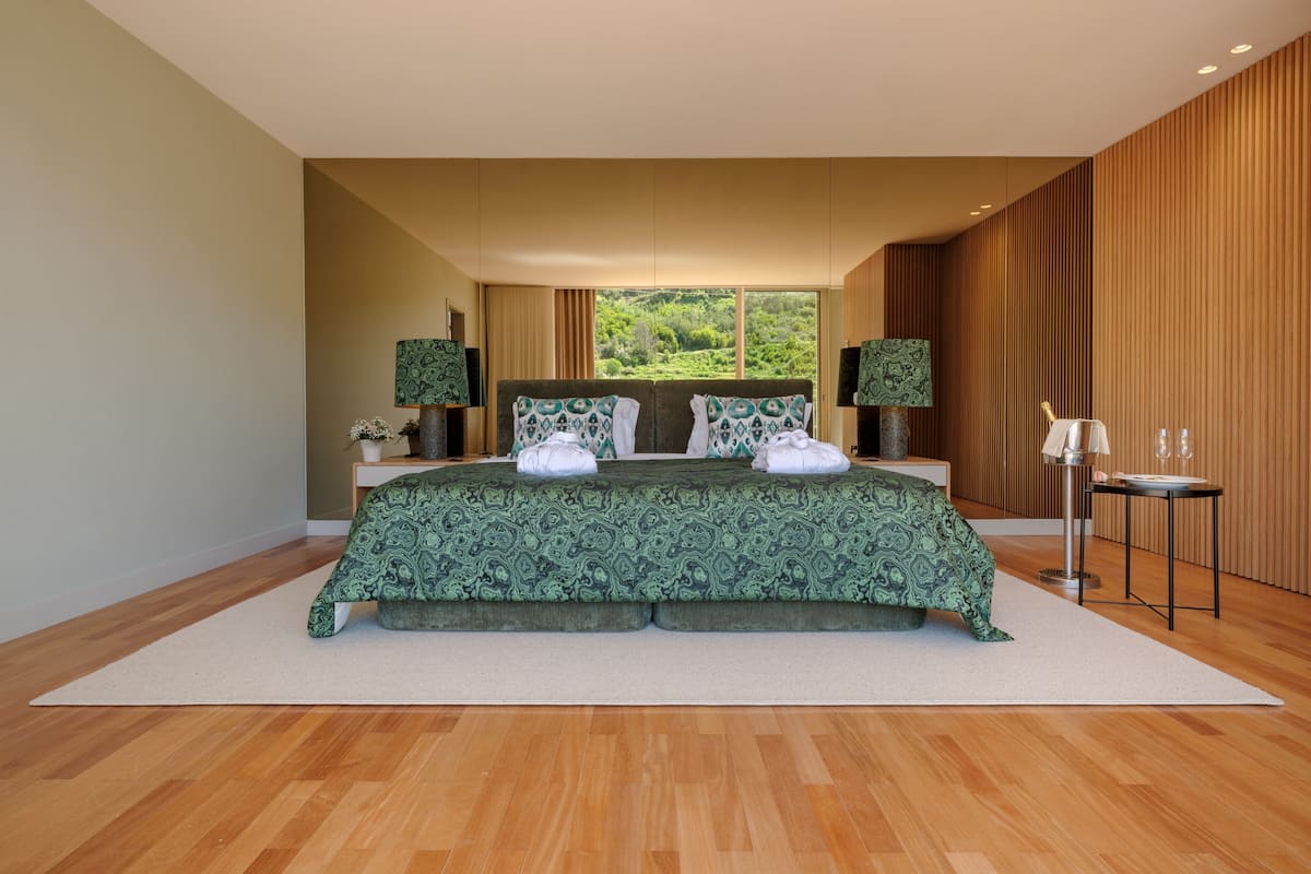 A modern bedroom with a large bed, two nightstands, a small table, and a view of greenery outside.