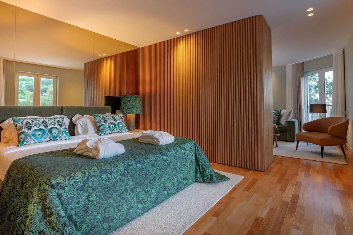 A modern bedroom with a large bed, a wooden accent wall, a sitting area with a chair, and a view of greenery outside.