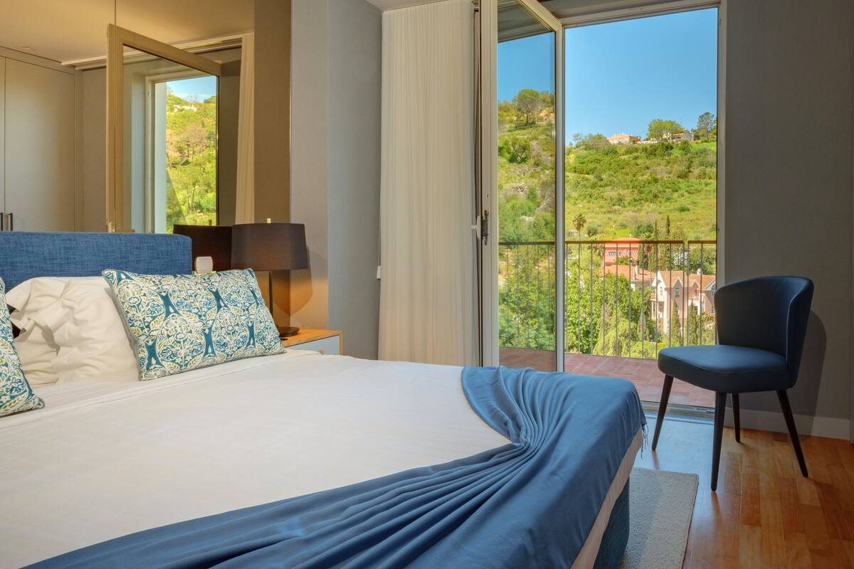 A bedroom with a bed, a chair, a nightstand, a lamp, and a balcony with a view of greenery and buildings.