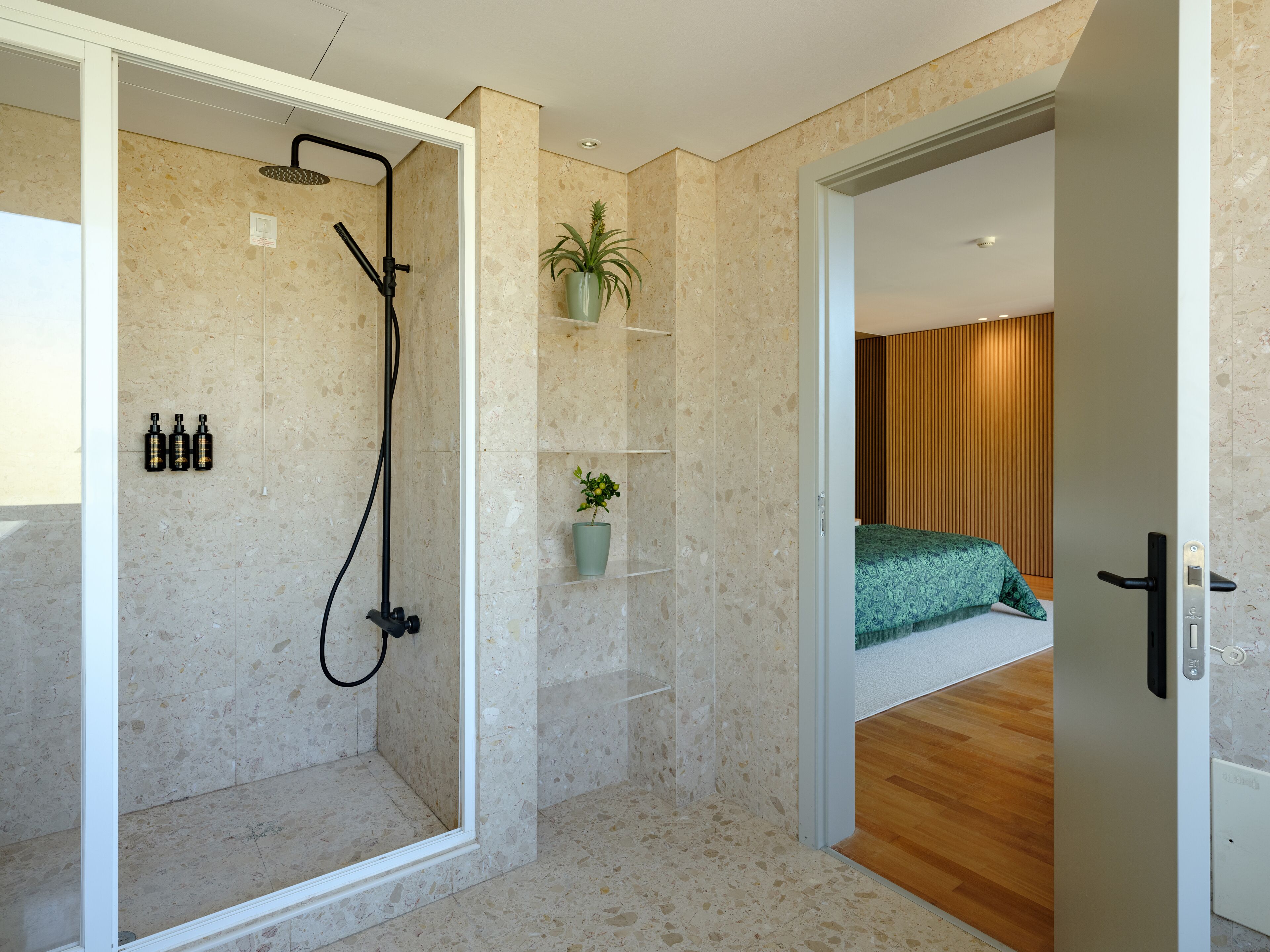 Un baño moderno con ducha, una planta en maceta y una cama visible a través de una puerta abierta.