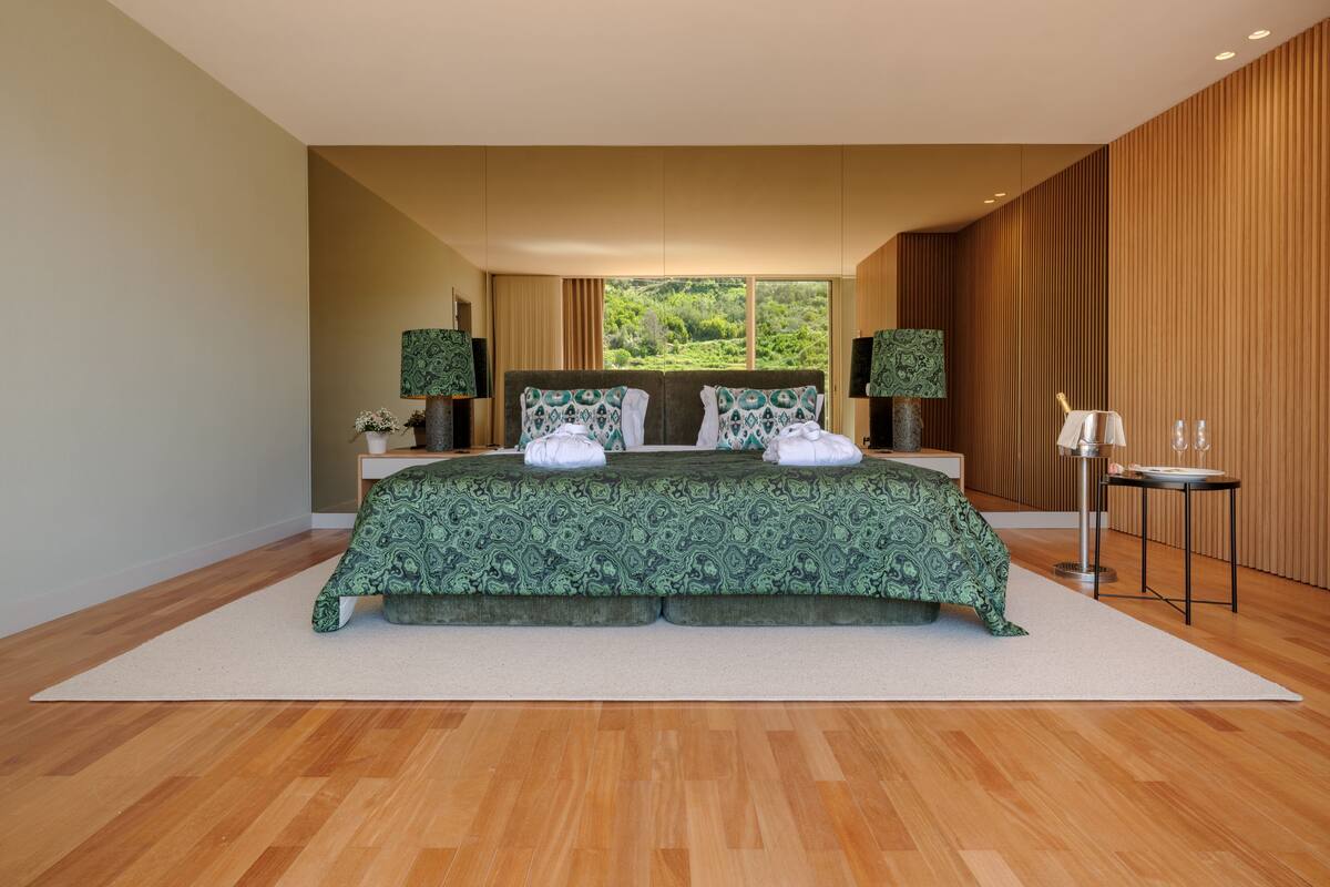 A modern bedroom with a large bed, two nightstands, a small table, and a view of greenery outside.