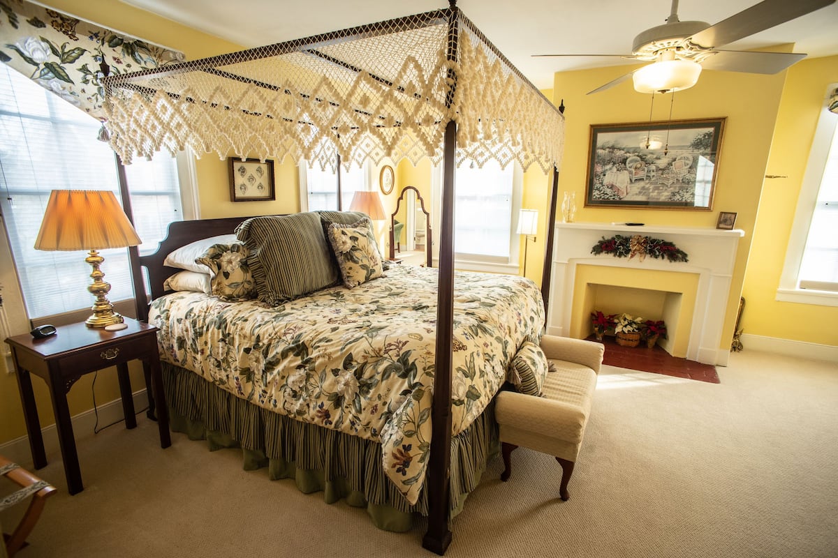 A four-poster bed with a floral bedspread, a nightstand with a lamp, a chair, and a fireplace with a mantel.