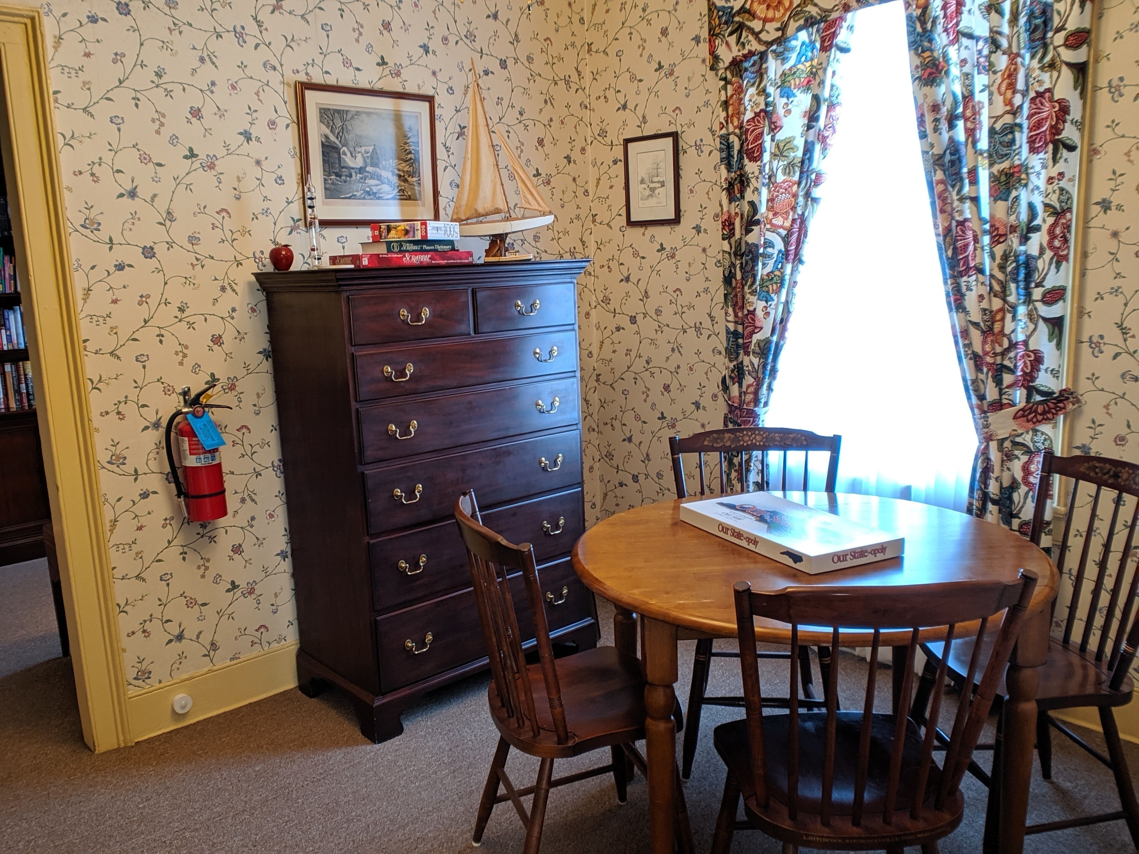 A dining area with a wooden table and chairs, a dark wooden chest of drawers, and a window with floral curtains.