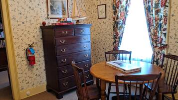 A dining area with a wooden table and chairs, a dark wooden chest of drawers, and a window with floral curtains.