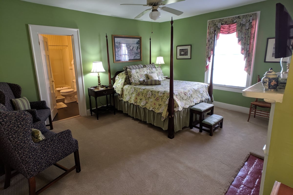 A bedroom with a four-poster bed, a chair, a nightstand, a bedside table, a window with curtains, and a ceiling fan.