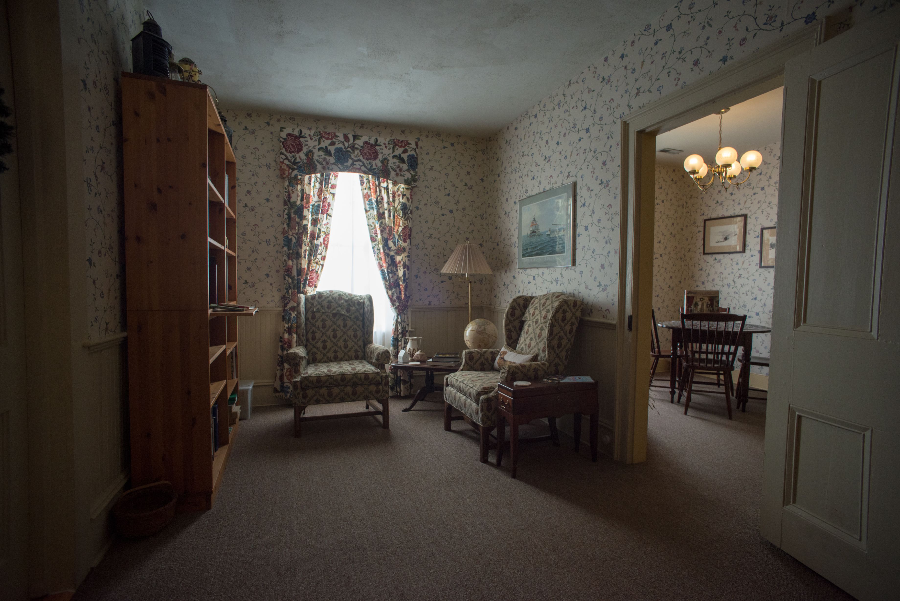 A cozy living room with a wooden bookshelf, patterned wallpaper, and a dining area with a chandelier.