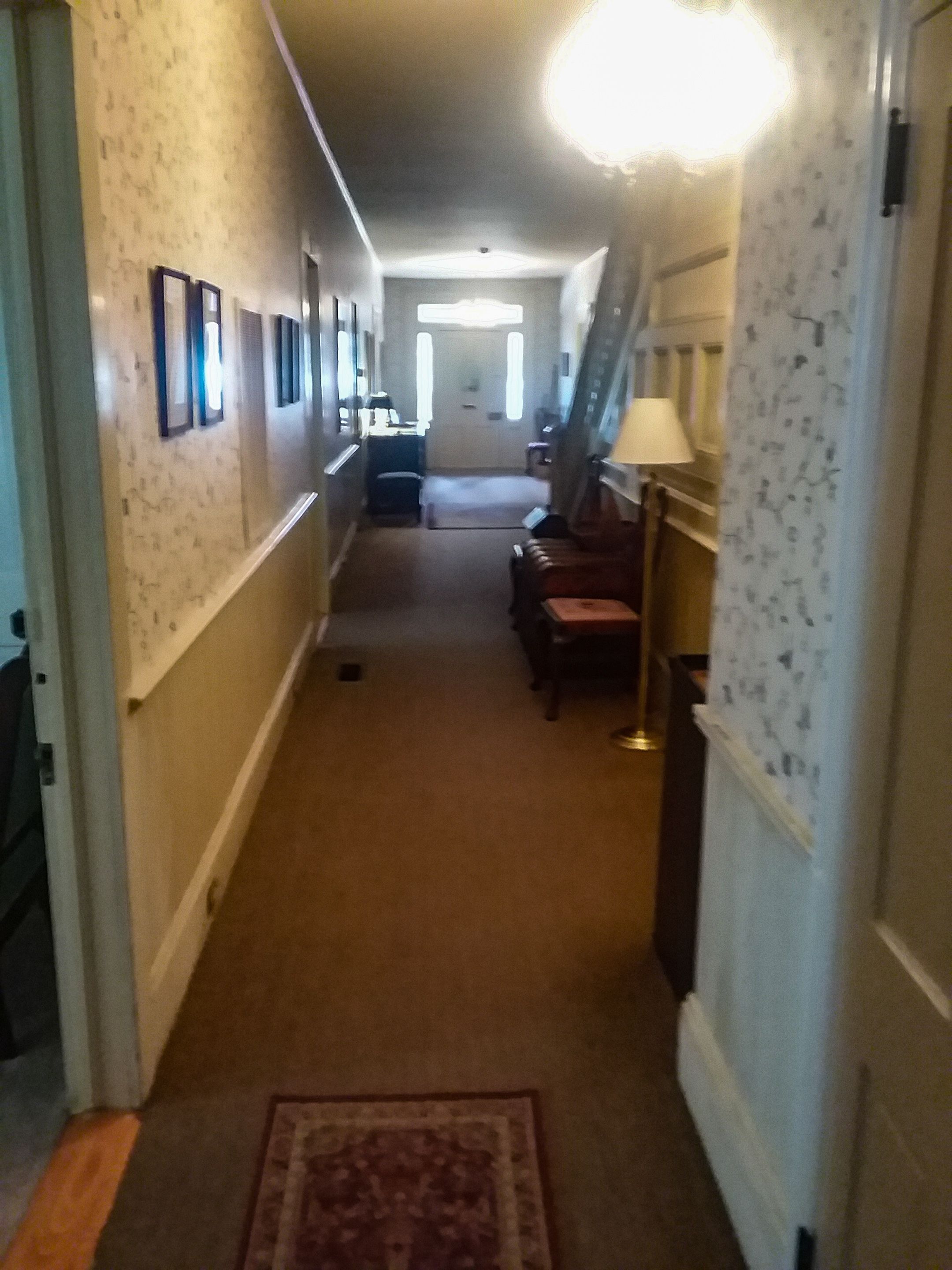 A hallway with a carpeted floor, framed pictures on the walls, a lamp, and a staircase.