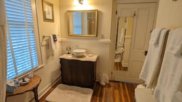 Un bany amb un tocador de fusta, un lavabo blanc, un mirall sobre el tocador i un vàter blanc. Hi ha un feser blanc a terra i un suport de tovalloles amb tovalloles.