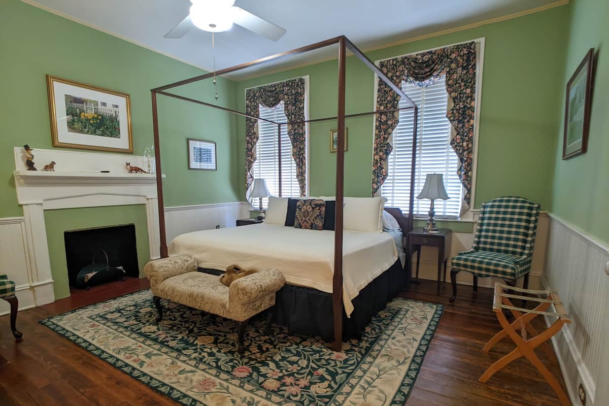 A bedroom with a four-poster bed, a fireplace, a green floral rug, and a checkered chair.