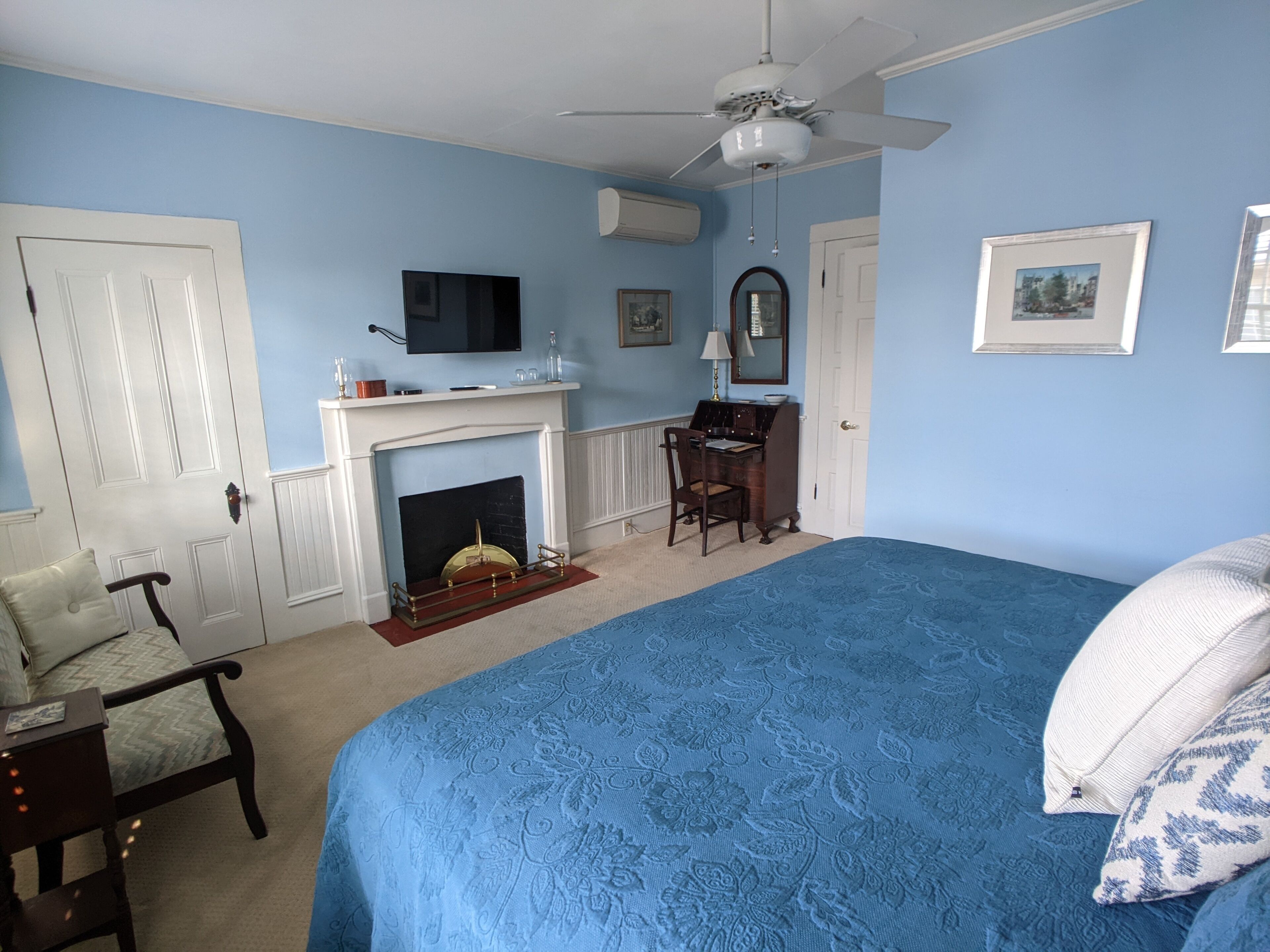 A bedroom with a large bed, a desk with a chair, a fireplace, a ceiling fan, and a television.