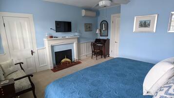 A bedroom with a large bed, a desk with a chair, a fireplace, a ceiling fan, and a television.