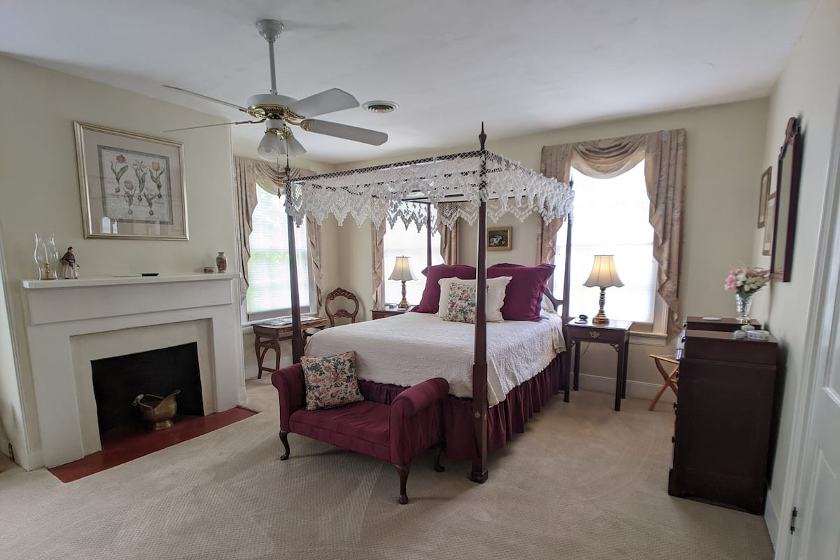 A four-poster canopy bed with a white bedspread and purple pillows, a fireplace with a mantel, and a small armchair.