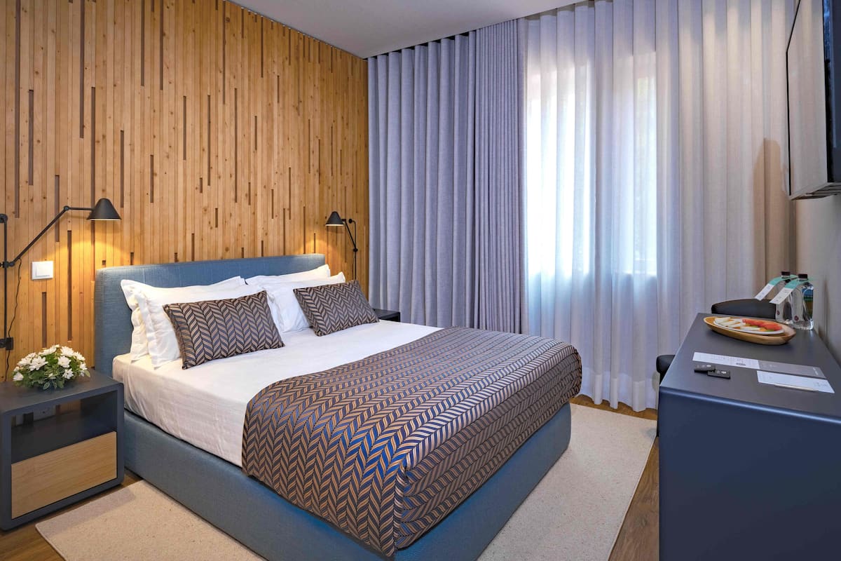 Un dormitorio moderno con un panel de madera en la pared, una cama grande con ropa de cama estampada, dos lámparas de noche y un escritorio con una computadora portátil y botellas de agua.
