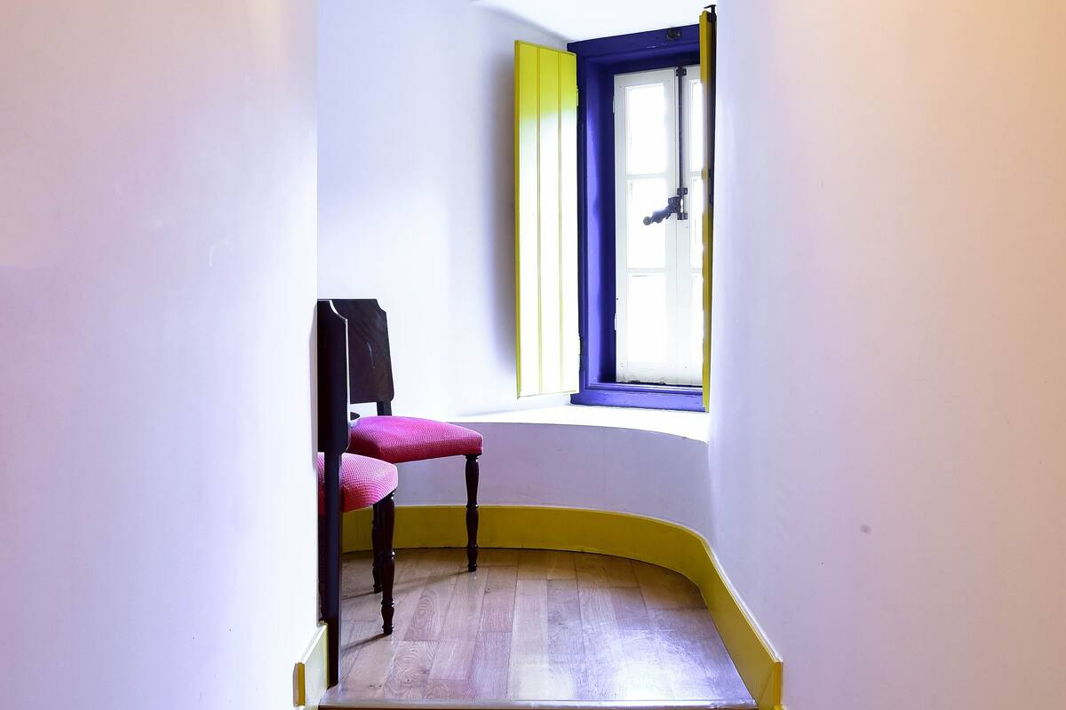 Un pasillo con piso de madera, una silla, un aplique en la pared y un ventanal con detalles azules y amarillos.