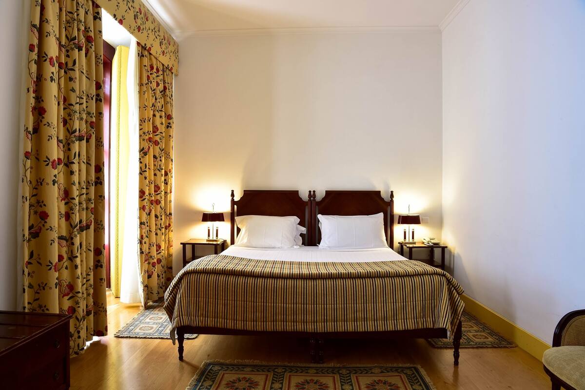 Habitación de hotel con una cama grande, dos mesitas de noche con lámparas, cortinas florales y suelo de madera.