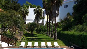 Piscina com espreguiçadeiras, um edifício branco com telhado vermelho e vegetação ao redor.