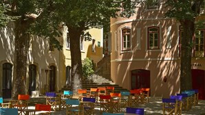 Uma área de estar ao ar livre com cadeiras e mesas de madeira, cercada por árvores e edifícios com janelas em arco e uma escadaria.