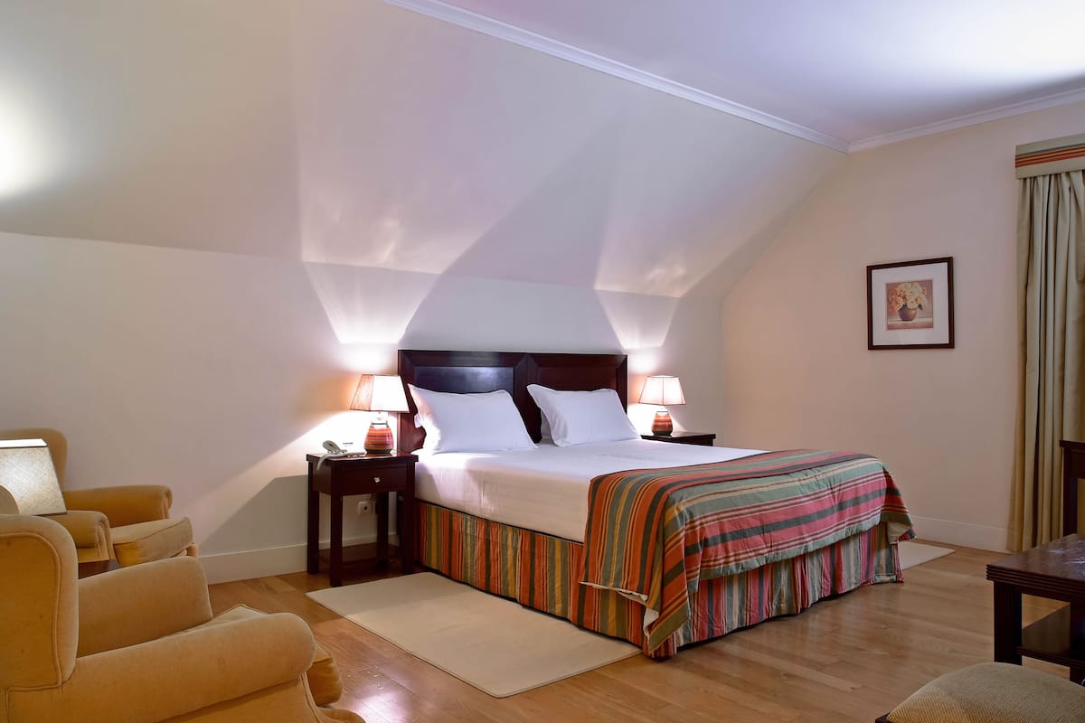 Un dormitorio con cama, lámparas de noche, una silla, una mesa y un cuadro en la pared.