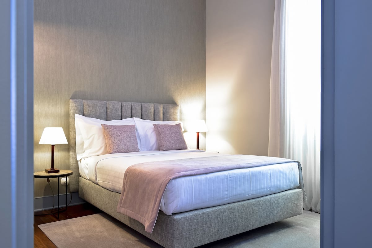 Una cama bien tendida con cabecera gris, dos lámparas de noche y una ventana con cortinas.