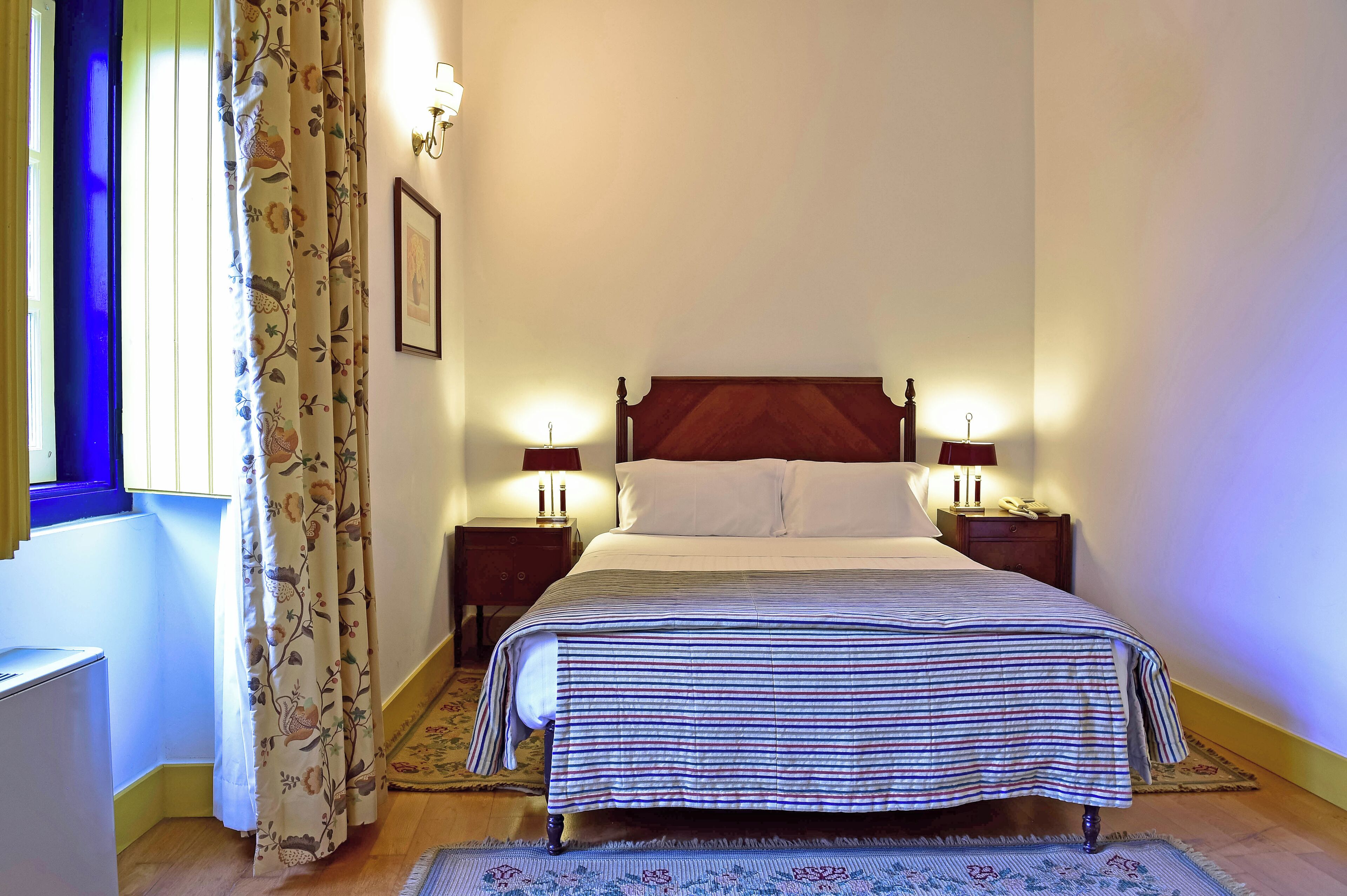 A neatly made bed with a striped bedspread, two bedside lamps, a nightstand, and a floral curtain.