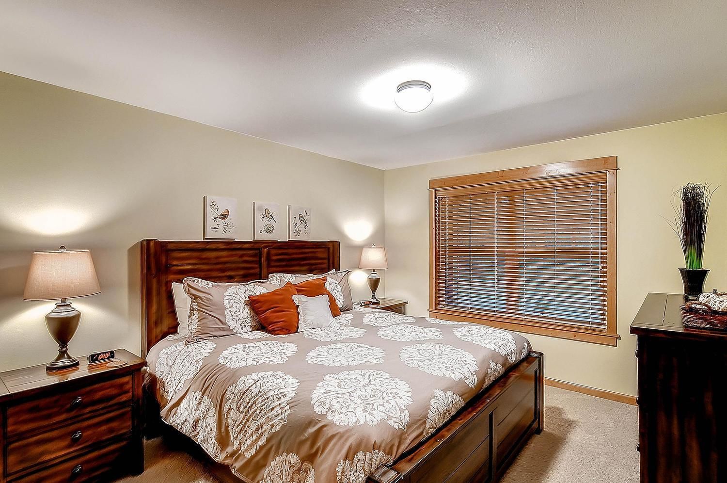A bedroom with a bed, two bedside tables, a nightstand, a window with blinds, and wall-mounted artwork.
