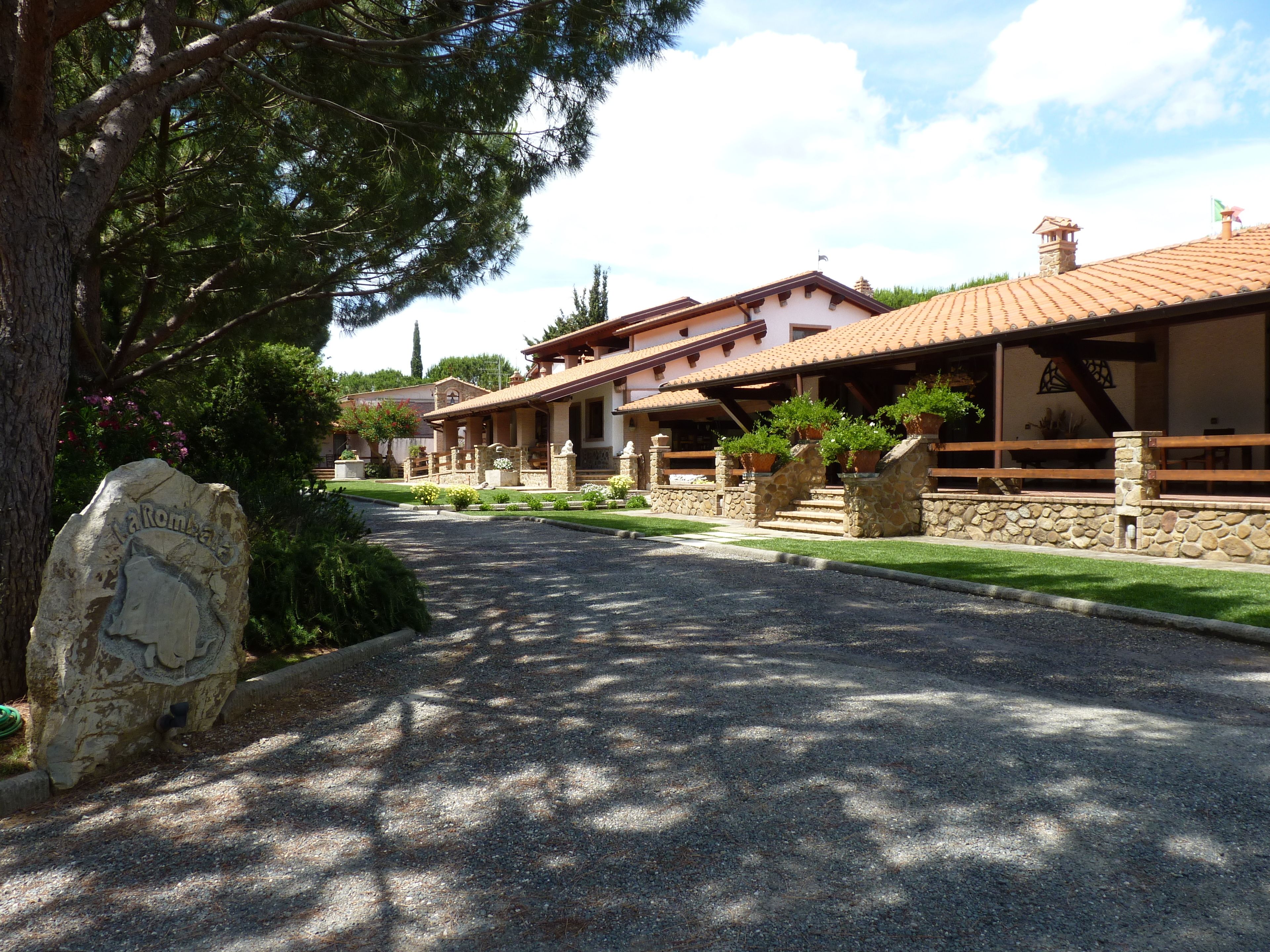 Un segnale in pietra raffigura una montagna e un cavallo, un vialetto sterrato conduce a un edificio con tetto in tegole e gradini in pietra, immerso nel verde.