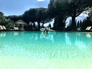 A clear pool with lounge chairs and a large tree in the background.