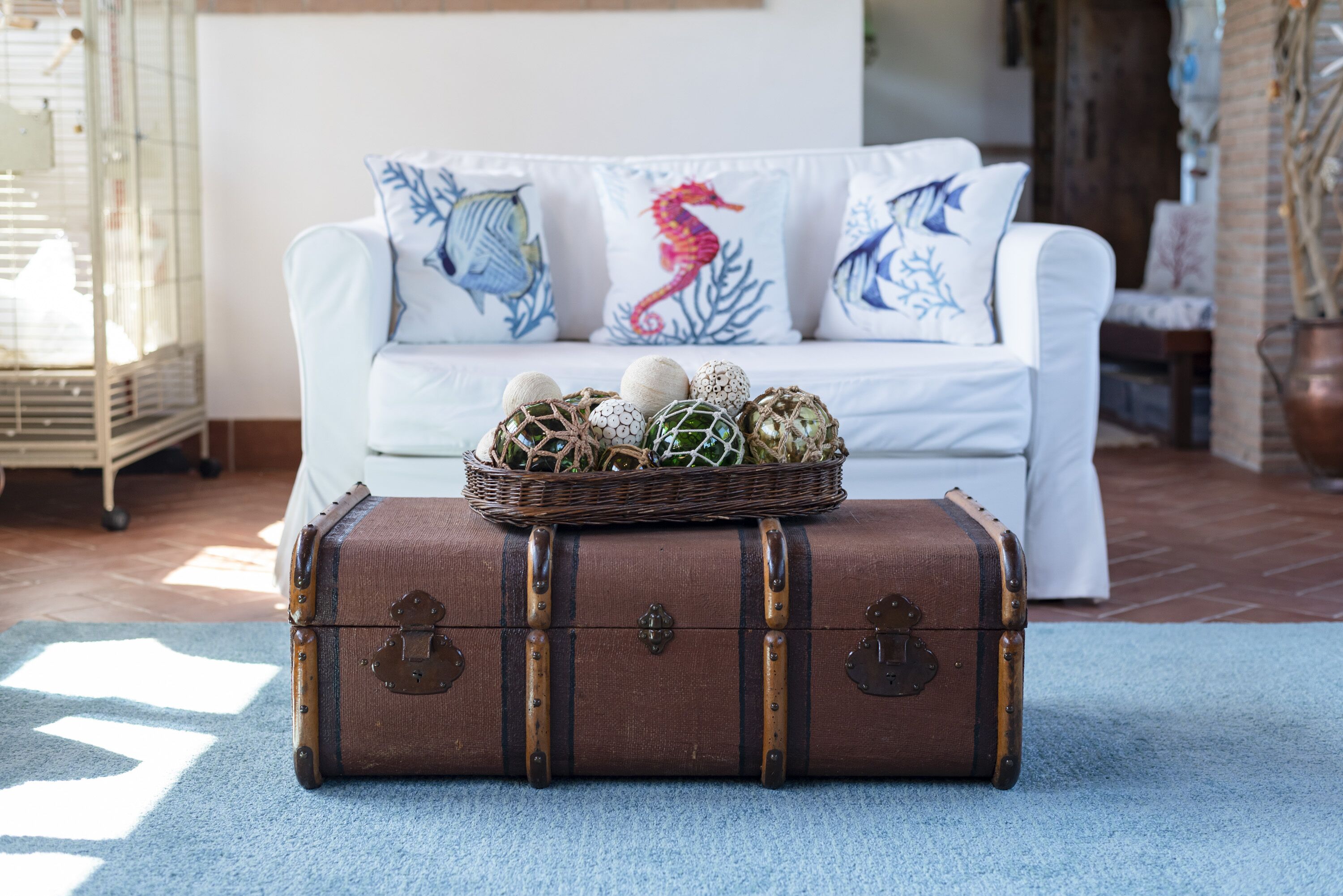 Ein alter Koffer mit Dekorationselementen auf einem blauen Teppich, ein weißes Sofa mit Seepferdchen-Kissen und ein Vogelkäfig im Hintergrund.