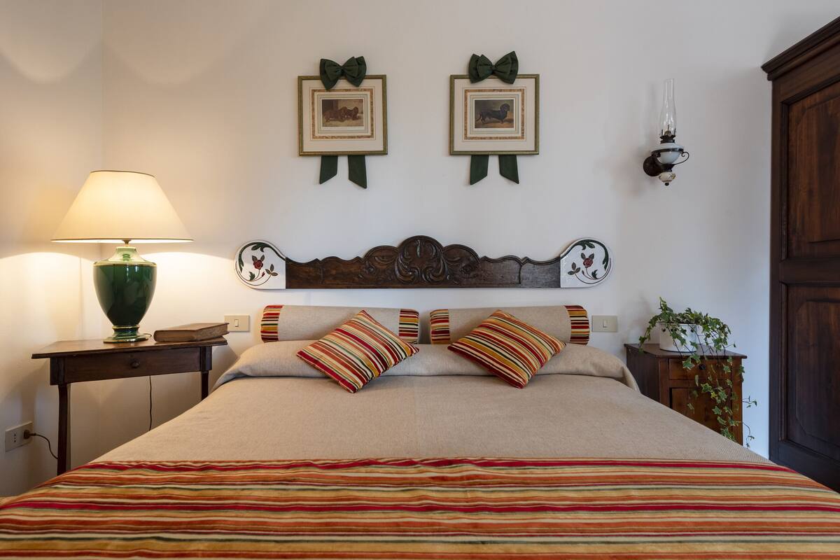 Een net opgemaakt bed met gestreepte lakens, een groene lamp en ingelijste foto’s aan de muur.
