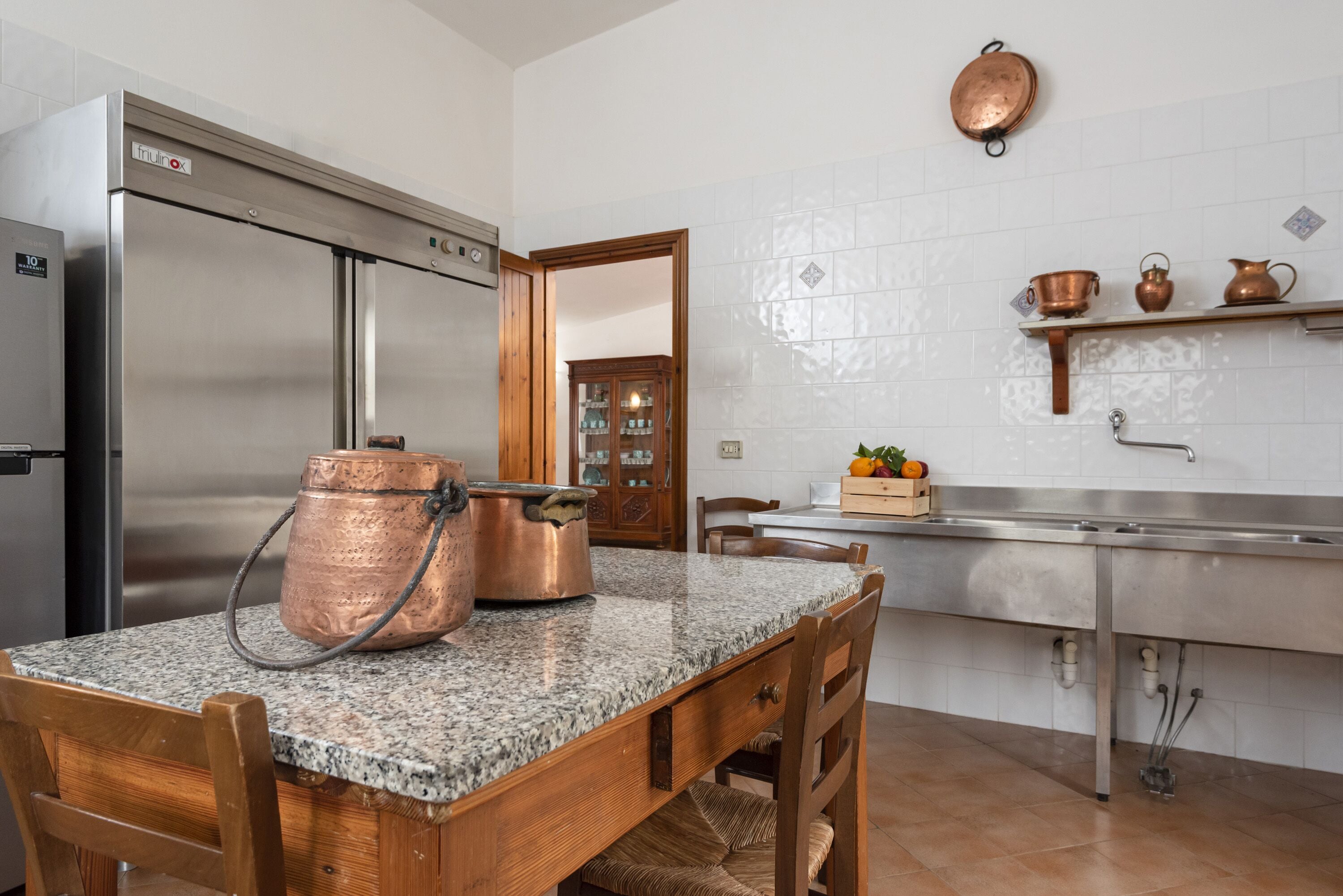 Una cucina con piani di lavoro in granito, pentole in rame, frigorifero in acciaio inossidabile e un tavolo da pranzo in legno con sedie.