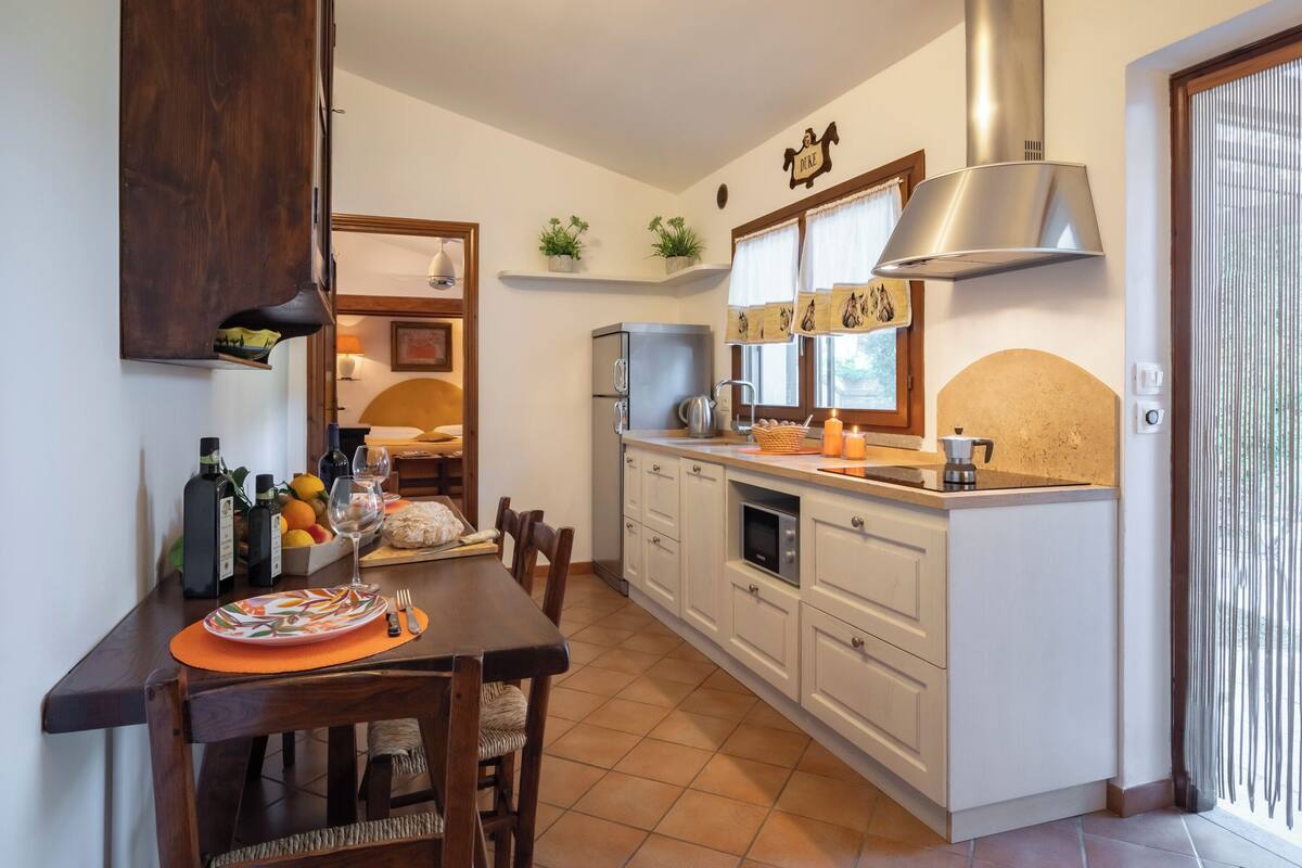 Een compacte keuken met witte kastjes, een ingebouwde oven, een roestvrijstalen afzuigkap en een eethoek met een houten tafel en stoelen.