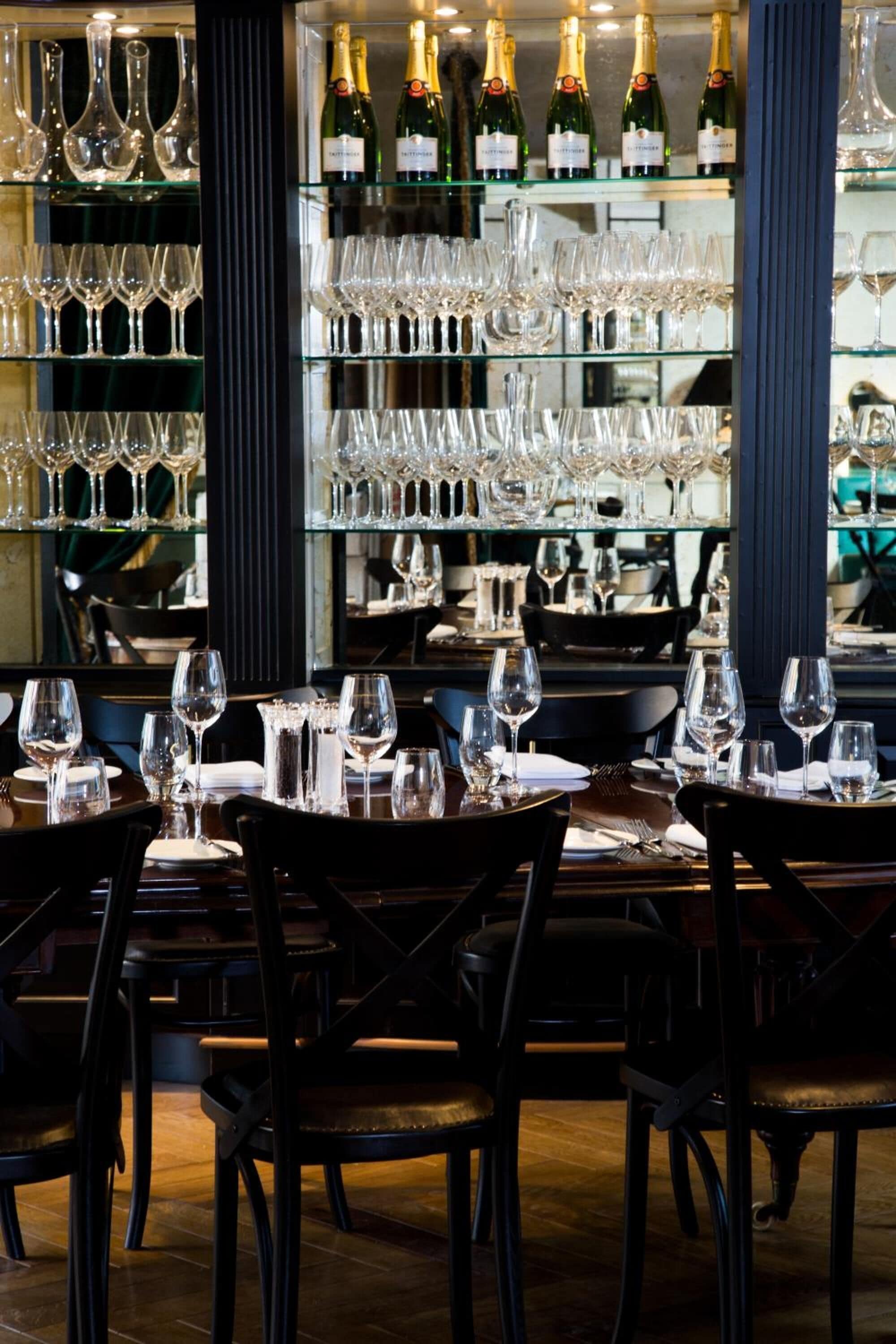 L’intérieur d’un restaurant présente de nombreuses tables dressées pour le service, un présentoir à vin contenant différentes bouteilles et un mur miroitant qui reflète l’espace réservé aux convives.
