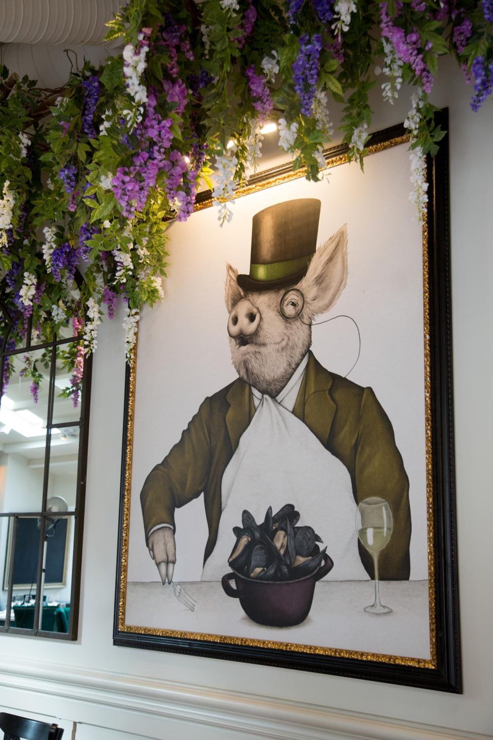 Une œuvre d’art encadrée représentant un cochon en costume et chapeau, un bol de champignons et un verre de vin.