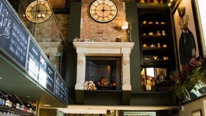 Un coin bar avec un mur en briques, une cheminée et une horloge. On y trouve des étagères remplies de bouteilles et un comptoir avec des verres.