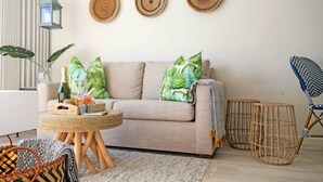 Una sala de estar con un sofá color beige, una mesa de centro redonda de madera, y una silla azul con estampado. Se observan tres adornos de pared tejidos sobre el sofá, una mesita auxiliar de mimbre y una lámpara de pie tejida.